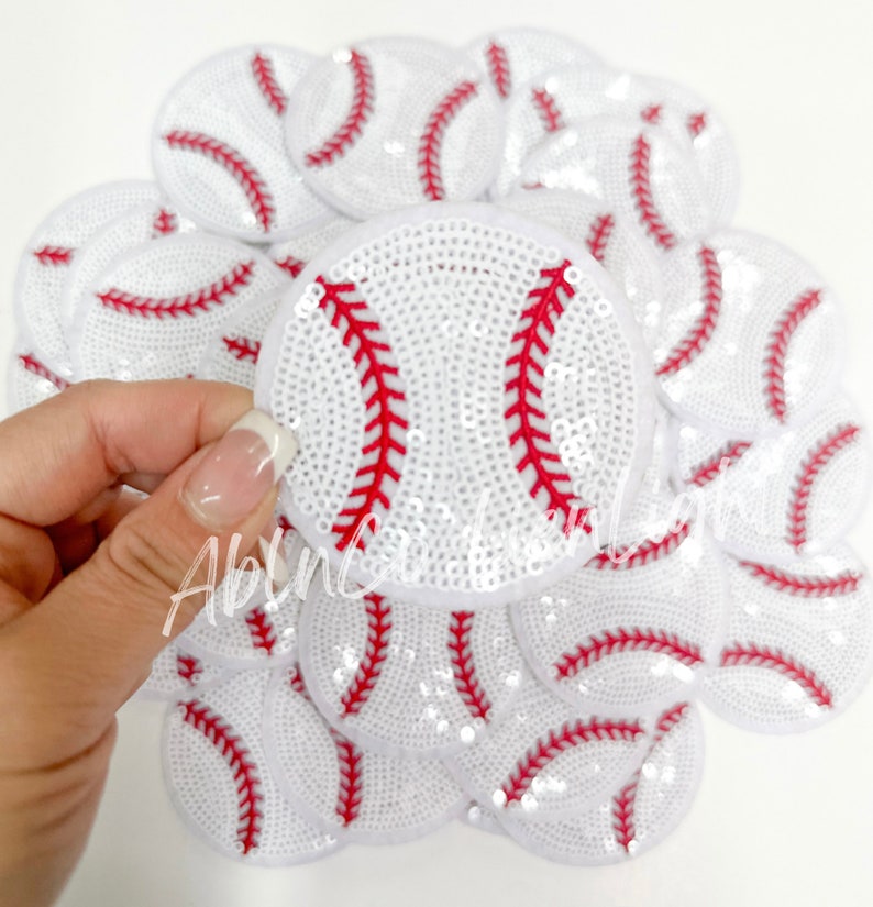 May include: A pile of white and red sequin baseball patches. The patches are approximately 5 cm in diameter and feature a white background with red stitching.