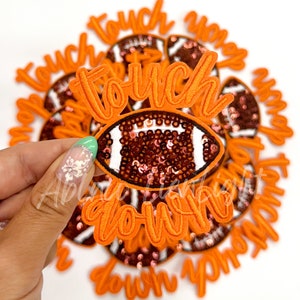 May include: A close-up of a hand holding an orange and brown felt patch with the words "touch down" and a sequined football design.