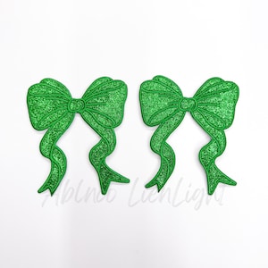 May include: Two green glitter bows with a shiny finish. The bows are made of fabric and have a loop in the center.