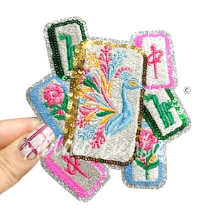 May include: Embroidered patch featuring a peacock with a colorful tail, surrounded by other embroidered elements. The patch has a silver glitter border and is held by a hand. The peacock is blue, and the surrounding elements are pink, green, and yellow.