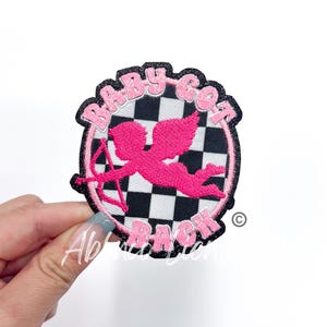 May include: Pink and black embroidered iron-on patch with a checkered background and a pink cherub with an arrow. The text "Baby Got Back" is written in a pink and black font around the edge of the patch.