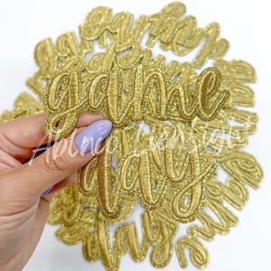 May include: Gold glitter fabric applique with the words "Game Day" in a cursive font. The applique is held in a hand with a light purple nail polish. Other appliques are in the background.