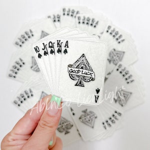May include: A stack of white felt patches with black embroidery of playing cards. The top patch is a black spade with the text "Good Luck" on a banner. The other patches are the 10, Jack, Queen, King, and Ace of spades.
