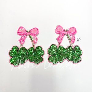 May include: Two green four-leaf clover patches with pink bows. The clovers are made of sequins and the bows are made of fabric.