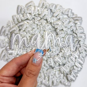 May include: Silver glitter iron-on letters spelling "mama" in a cursive font. The letters are white with a silver glitter outline.