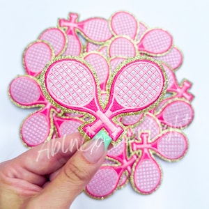 May include: A pile of pink and gold glitter iron-on patches shaped like tennis rackets. The patches are approximately 2 inches in diameter.