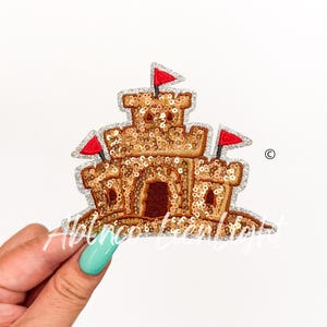 May include: A sparkly gold sandcastle with red flags on top. The sandcastle is on a white background.