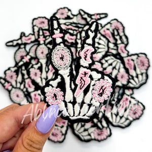 May include: A pile of black and white skeleton hand patches with pink glitter accents. The patches are embroidered and have a punk rock aesthetic.