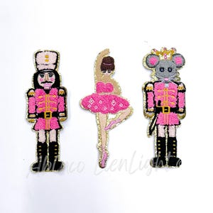 May include: Three pink and gold embroidered felt Christmas ornaments. The ornaments are a ballerina, a nutcracker, and a mouse king.