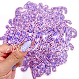 May include: A pile of purple and white tie-dye iron-on patches with the word "wifey" in a glittery font.
