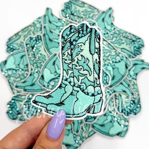 May include: A collection of turquoise cowboy boot stickers. The boots have a detailed floral design and are embellished with small, sparkling gems. The stickers are a light teal color with a white border.