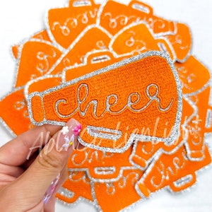 May include: Orange felt iron-on patch with silver glitter trim shaped like a megaphone with the word "cheer" embroidered in silver glitter thread.