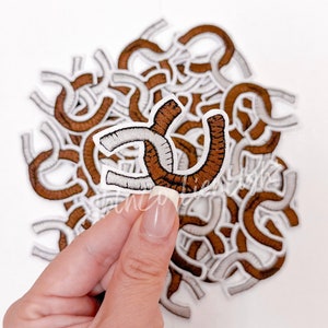 May include: A pile of brown and white horseshoe-shaped fabric patches. The patches are embroidered with a white outline and brown stitching.