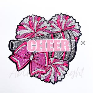 May include: A pink and silver sequin heart-shaped cheerleading patch with a megaphone, pom-poms, and a bow. The word "CHEER" is written in pink letters on the megaphone.