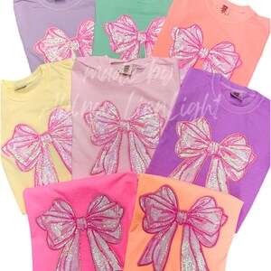 May include: Seven t-shirts in various pastel colors with a large pink sequined bow design on the front. The bows are all the same design but are on different colored shirts.