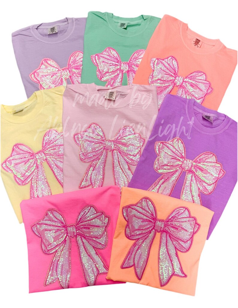 May include: Seven t-shirts in various pastel colors with a large pink sequined bow design on the front. The bows are all the same design but are on different colored shirts.