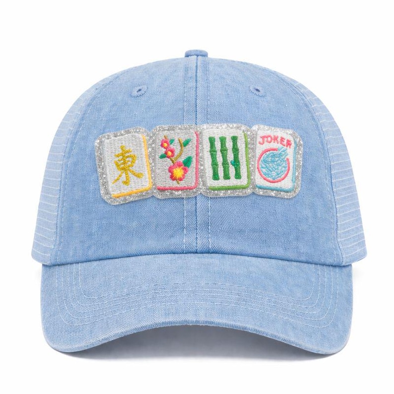 K&ouml;nnte beinhalten: Hellblaue Jeans-Baseballkappe mit Mesh-R&uuml;ckseite. Auf der Vorderseite befinden sich vier gestickte Aufn&auml;her mit chinesischen Schriftzeichen, Blumen, Bambus und dem Wort "JOKER". Die Aufn&auml;her sind mit silbernem Glitzer umrandet.