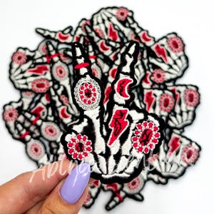 May include: A pile of black and white skeleton hand patches with red and white details. The patches feature a skeleton hand making a peace sign with a lightning bolt and crescent moon design.