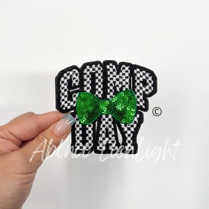 May include: A black and white checkered iron-on patch with the text "COMP DAY" and a green sequin bow tie.