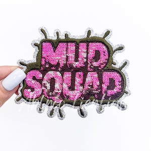May include: A pink sequined patch with the words "MUD SQUAD" in a bubble font. The patch has a dark green border and a silver glitter outline. The patch is on a white background.