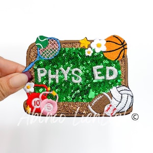 May include: A brown and gold embroidered patch with a green field of sequins. The patch says "PHYS ED" in white letters. The field is surrounded by various sports equipment, including a basketball, a volleyball, a tennis racket, a dumbbell, and a football.