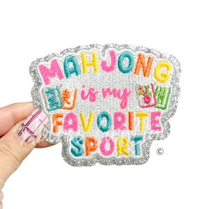 May include: Embroidered patch with the text "MAHJONG is my FAVORITE SPORT" in colorful letters. The patch has a silver glitter border and features images of Mahjong tiles. The patch is held by a hand.