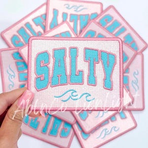 May include: A stack of pink and white embroidered iron-on patches with the word "SALTY" in blue lettering and a wave design.