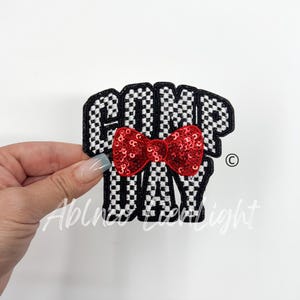 May include: Black and white checkered fabric patch with the words "Camp Day" in white lettering. A red sequined bow is centered on the patch.