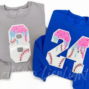 May include: Two sweatshirts, one gray and one blue, each with a large number applique. The appliques feature a baseball design with pink, blue, and white accents. The gray sweatshirt has the number 8, and the blue sweatshirt has the number 24.