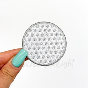 May include: A white golf ball patch with a silver outline and a dimpled surface.
