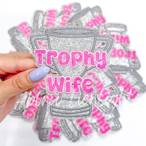 May include: A silver glitter trophy with the words "Trophy Wife" in pink glitter lettering. The trophy is surrounded by other similar trophies with different text.