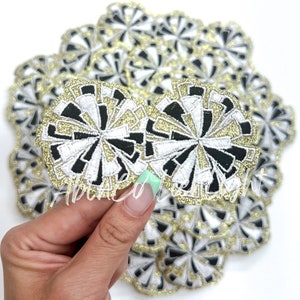 May include: A pile of black, white, and gold glitter iron-on patches in the shape of a stylized flower. The patches are approximately 2 inches in diameter.