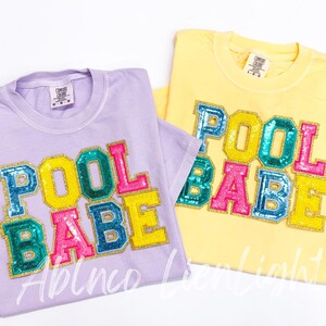 May include: Two t-shirts with the words "Pool Babe" in colorful, glittery letters. The shirts are in lavender and yellow.