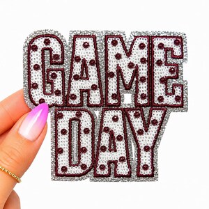 May include: A decorative patch featuring the text GAME DAY in block letters. The letters are filled with white sequins and accented with small maroon circular studs. The entire design is outlined with a thick, shimmering silver border.