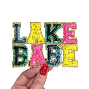 May include: A colorful sequined patch with the words "LAKE BABE" in bold, block letters. The letters are in shades of yellow, pink, teal, and black, outlined with gold glitter. The patch is held by a hand with red nail polish.