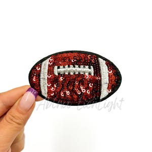 May include: A red and white football-shaped patch with sequins. The football has a white stitching design and is surrounded by a black border.