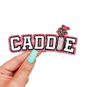 May include: Pink glitter iron-on patch with the word "CADDIE" in white letters with black outlines. A golf bag with clubs and a flag is on the right side of the patch.