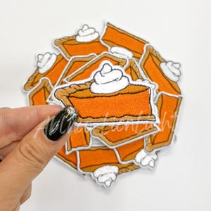 May include: Embroidered patches featuring slices of pumpkin pie with whipped cream. The patches are orange, brown, and white, with detailed stitching. A hand holds one patch, with others scattered in the background. The patches are likely for clothing or accessories.