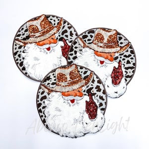 May include: Three round Christmas ornaments featuring Santa Claus in a cowboy hat. The hats have a brown and gold pattern, and the ornaments have a black and white cow print background. Santa has a white beard and is giving a hand gesture.