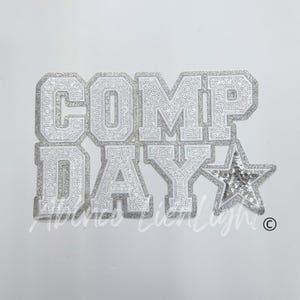 May include: A silver and white sequined patch with the words "COMP DAY" in a collegiate font. A sequined star is to the right of the text. The patch is on a white background.