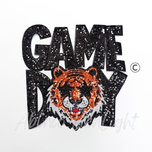 May include: A black sequined patch with the words "GAME DAY" and a tiger face. The tiger has orange and white stripes, a red mouth, and is centered below the text. The patch is on a white background.