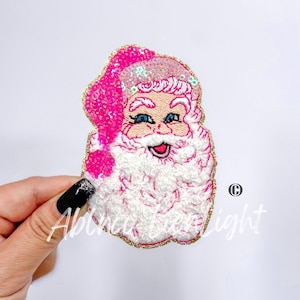 May include: Pink and white embroidered Santa Claus patch with a gold glitter outline. The Santa has a white beard and a pink hat with sequins.