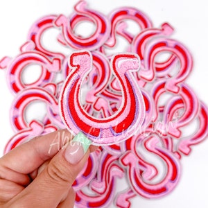 May include: A pile of pink and red horseshoe-shaped iron-on patches. The patches have a white outline and are arranged in a circle.