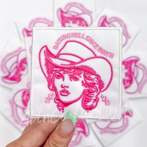May include: A white embroidered patch with a pink outline of a woman wearing a cowboy hat. The text "Raising Hell Since Birth" is written around the woman's head.