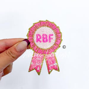 May include: A pink embroidered patch in the shape of a ribbon with the letters "RBF" in the center. The patch has a gold border and sequined ribbon tails. The patch is held by a hand.
