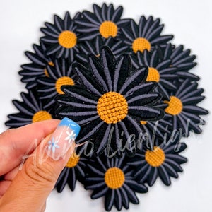 May include: A pile of black and gray felt daisy patches with a yellow center. The patches are arranged in a circle with one patch being held in the center by a hand.