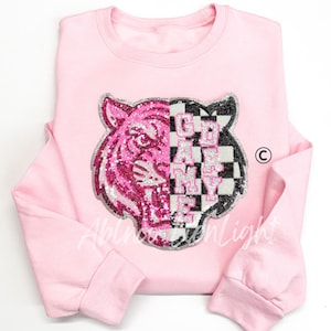 May include: A light pink sweatshirt featuring a sequined tiger face design. The tiger's face is split, with one half in pink sequins and the other in a black and white checkerboard pattern with the text "GAME MY LIFE".