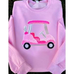May include: Pink sweatshirt with an embroidered pink golf cart design. The golf cart has silver golf clubs and a steering wheel.