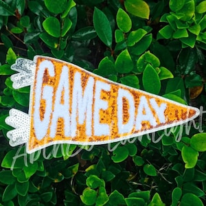 May include: A gold and white felt pennant with the text "GAME DAY" in white letters. The pennant is decorated with sequins.