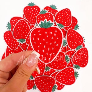 May include: A pile of red embroidered strawberry patches with white outlines and green leaves. The patches are arranged in a circle with one patch being lifted up by a hand.